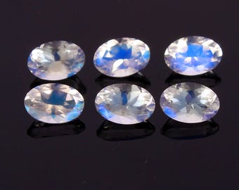May include: Six oval-cut moonstones arranged in two rows of three. Each gemstone features a translucent, clear body with a distinct blue adularescence or flash visible across the faceted surfaces, reflecting light against a dark background.