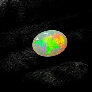 May include: An oval-shaped cabochon opal gemstone featuring a translucent white base with vibrant, multi-colored play-of-color flashes in bright neon green, yellow, and orange hues distributed across the surface in a distinct honeycomb pattern.