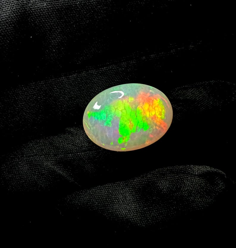 May include: An oval-shaped cabochon opal gemstone featuring a translucent white base with vibrant, multi-colored play-of-color flashes in bright neon green, yellow, and orange hues distributed across the surface in a distinct honeycomb pattern.