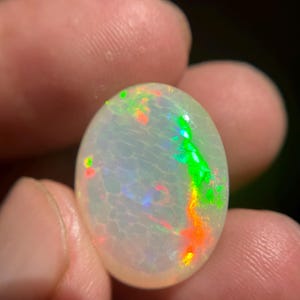 May include: An oval-shaped cabochon opal gemstone featuring a translucent white base with a distinct honeycomb pattern. The stone displays vibrant flashes of neon green and bright orange play-of-color across its polished, smooth, and rounded surface.