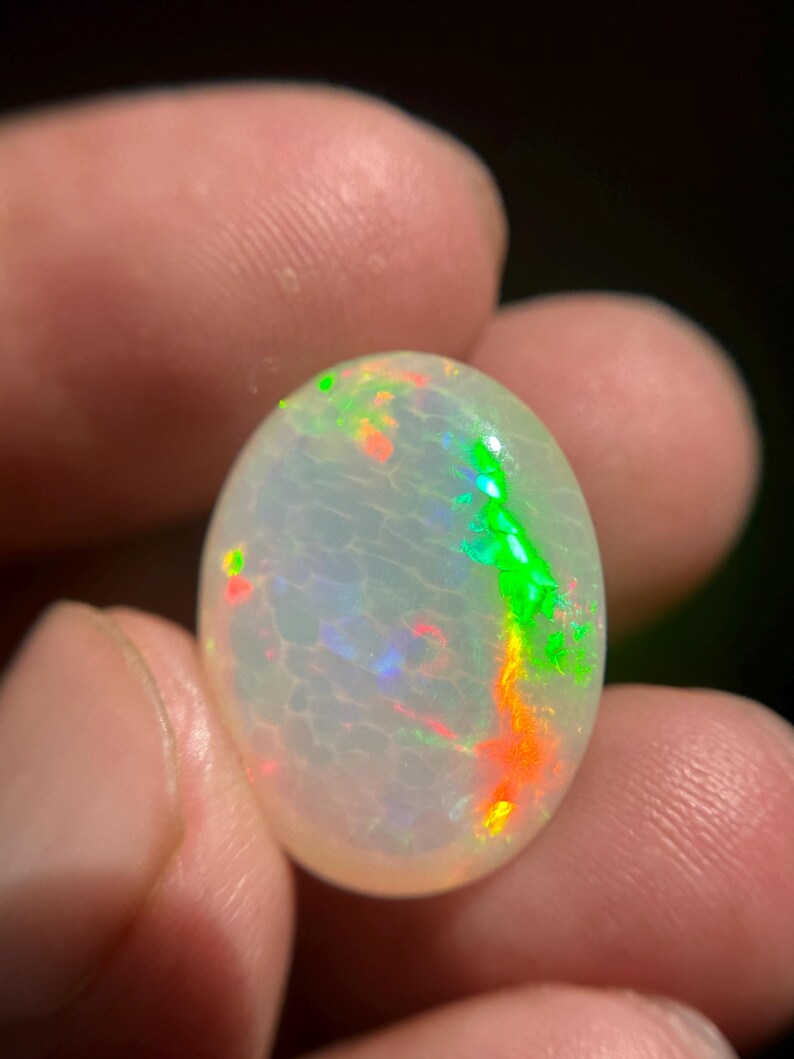 May include: An oval-shaped cabochon opal gemstone featuring a translucent white base with a distinct honeycomb pattern. The stone displays vibrant flashes of neon green and bright orange play-of-color across its polished, smooth, and rounded surface.