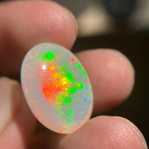 May include: An oval-shaped white opal gemstone featuring a translucent body with vibrant flashes of red, orange, and green play-of-color throughout its surface. The stone has a smooth, polished cabochon cut that reflects light across its rounded top.