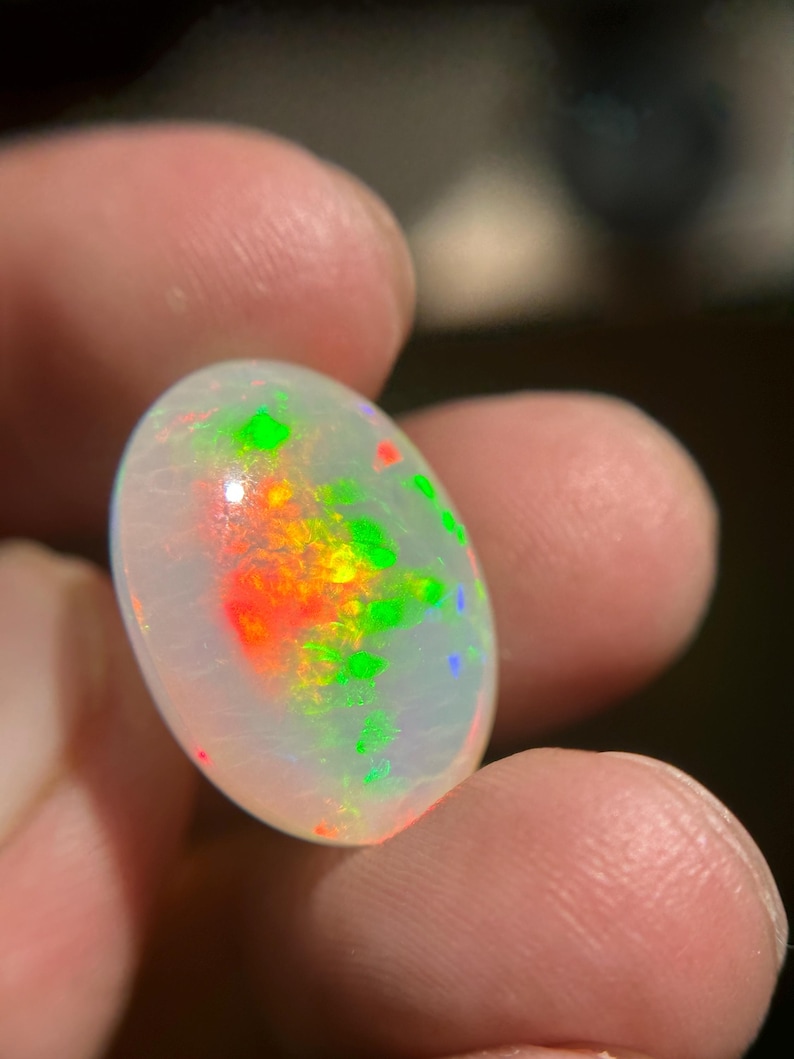 May include: An oval-shaped white opal gemstone featuring a translucent body with vibrant flashes of red, orange, and green play-of-color throughout its surface. The stone has a smooth, polished cabochon cut that reflects light across its rounded top.