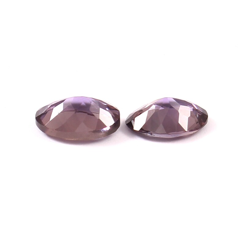 May include: Two oval-shaped, faceted gemstones with a translucent, light purple hue. Each stone features a polished surface with multiple geometric facets that reflect light, creating a shimmering appearance against a plain white background.