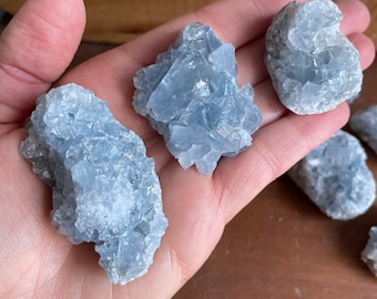 May include: Four blue celestite crystal clusters of varying sizes. The crystals are a light blue color and have a rough, textured surface.