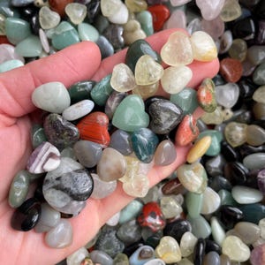 May include: A handful of polished gemstones in various colors, including green, red, gray, and yellow. Some stones are heart-shaped. The gemstones are smooth and appear to be tumbled. The image is a close-up shot of the stones.