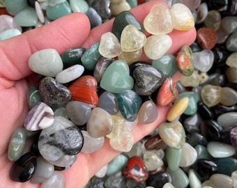 May include: A handful of polished gemstones in various colors, including green, red, gray, and yellow. Some stones are heart-shaped. The gemstones are smooth and appear to be tumbled. The image is a close-up shot of the stones.