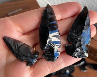 May include: Three black obsidian arrowheads with white swirls and patterns. The arrowheads are sharp and have a pointed tip.