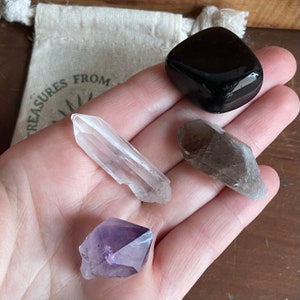 May include: A collection of four crystals: a clear quartz point, a smoky quartz point, a purple amethyst point, and a black obsidian stone.