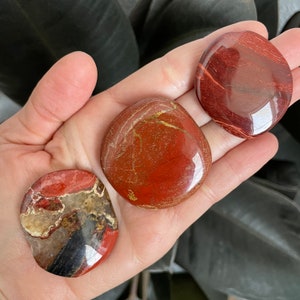 Pu&ograve; includere: Tre pietre di diaspro rosso, ciascuna con un motivo unico di rosso, marrone e oro. Le pietre sono lisce e levigate e sono disposte in una mano.