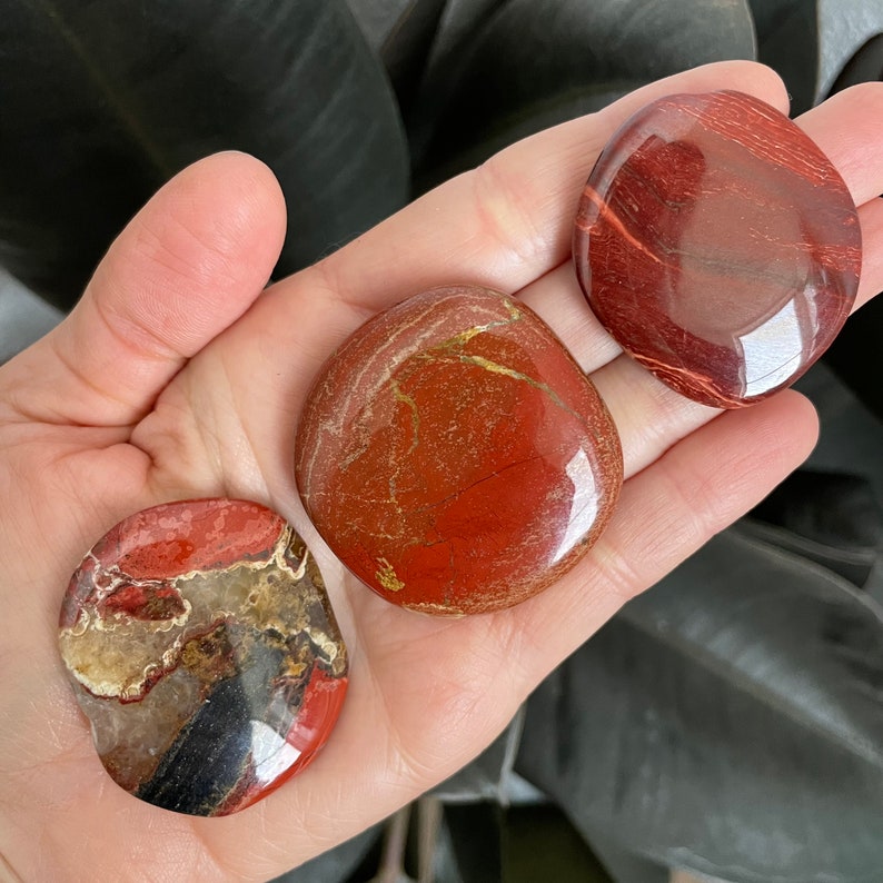 Pu&ograve; includere: Tre pietre di diaspro rosso, ciascuna con un motivo unico di rosso, marrone e oro. Le pietre sono lisce e levigate e sono disposte in una mano.