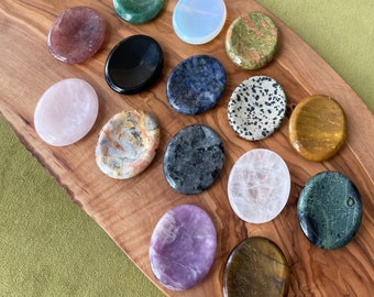 May include: A collection of polished, oval-shaped gemstones in various colors, including pink, black, green, and speckled patterns. The stones are arranged on a wooden board, showcasing their natural textures and hues. The gemstones are a mix of different types.