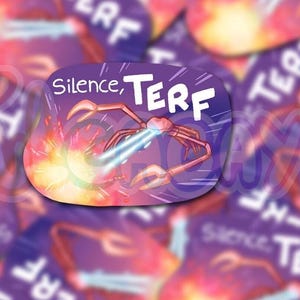 May include: A sticker featuring the words "Silence, TERF" in white on a purple background. A cartoon crab fires a laser beam. The design includes fiery, colorful elements.