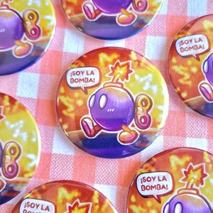 Puede incluir: Un conjunto de botones redondos y coloridos con un personaje de bomba de dibujos animados. La bomba es morada con una mecha encendida y el texto "¡SOY LA BOMBA!" en un bocadillo. El fondo es un degradado de amarillo y naranja.