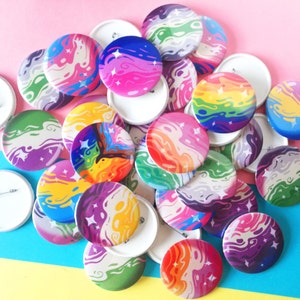 Puede incluir: Un conjunto de botones redondos y coloridos con diseños abstractos y arremolinados en varios colores, incluyendo rosa, azul, morado, verde y naranja. Algunos botones tienen detalles de estrellas. Los botones están dispersos sobre una superficie rosa y azul.