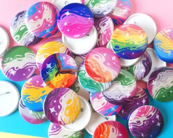 May include: A collection of colorful, round pinback buttons with swirling, abstract designs in various colors, including pink, blue, purple, green, and orange. Some buttons feature star accents. The buttons are scattered on a pink and blue surface.