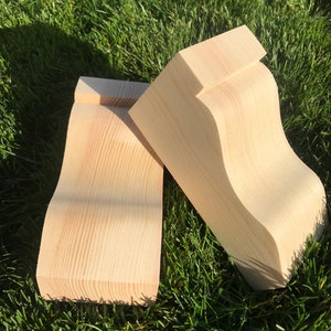 May include: Three unfinished wooden corbels with a curved design. The corbels are made of light-coloured wood and are positioned on a bed of green grass. The corbels have a smooth, natural finish, showcasing the wood grain.