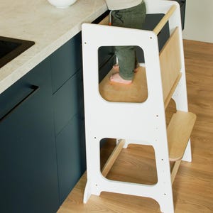 May include: A white wooden kitchen step stool with a natural wood platform and step. The step stool has a rectangular frame with cut-out sections. A child is standing on the platform near a countertop with a bowl of fruit.