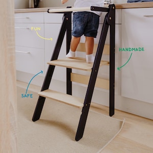 May include: A black and natural wood step stool with three steps. The stool is designed for children to reach higher surfaces safely. The words "FUN", "SAFE", and "HANDMADE" are written on the image.