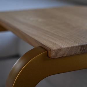 Op de afbeelding: Close-up van een houten tafel met een lichtbruin, rechthoekig tafelblad. De tafel heeft een gebogen, goudkleurig metalen onderstel. De houtnerf is zichtbaar op het tafelblad. De tafel heeft een modern design.