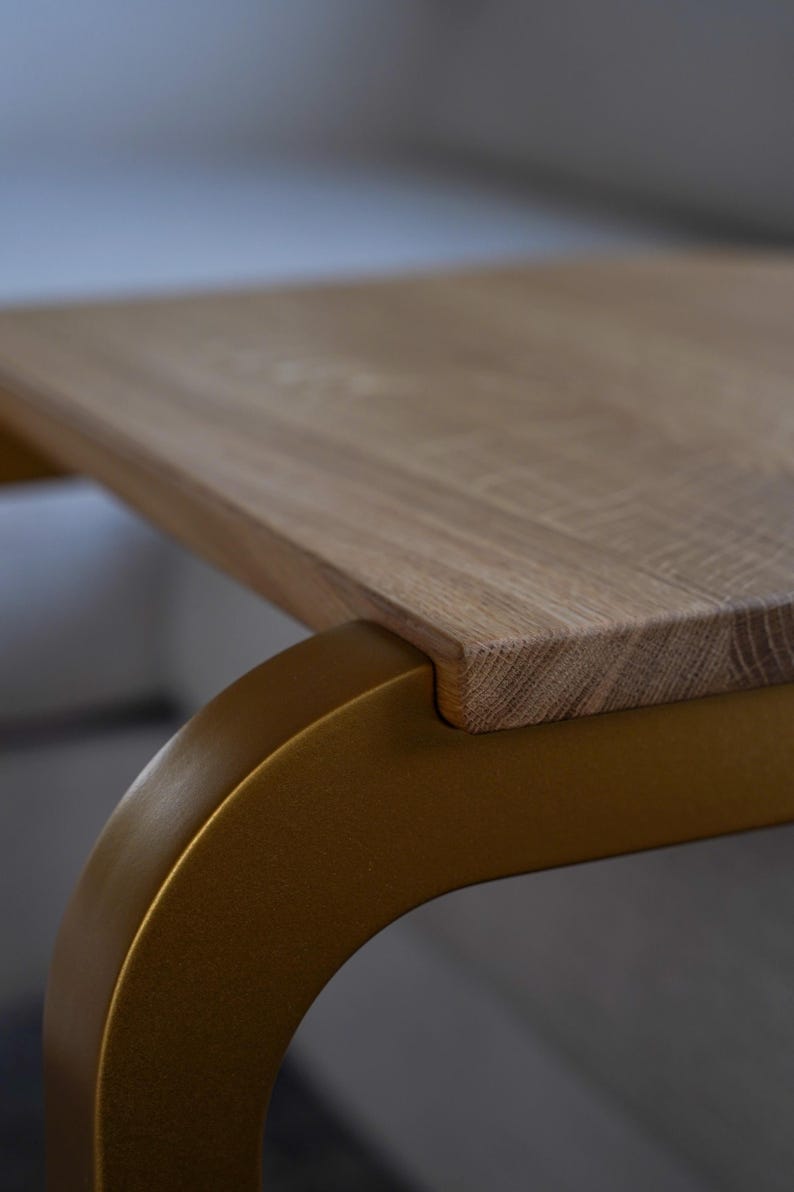 Op de afbeelding: Close-up van een houten tafel met een lichtbruin, rechthoekig tafelblad. De tafel heeft een gebogen, goudkleurig metalen onderstel. De houtnerf is zichtbaar op het tafelblad. De tafel heeft een modern design.