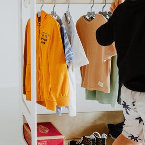 May include: A white wooden clothing rack with a shelf on top. The rack is filled with children's clothing, including a yellow hoodie with the text "it's all good", a white t-shirt, a blue and white patterned shirt, and a tan t-shirt. There is a pair of leopard print Vans shoes on the bottom shelf, along with a red box that says "Vans" on it.