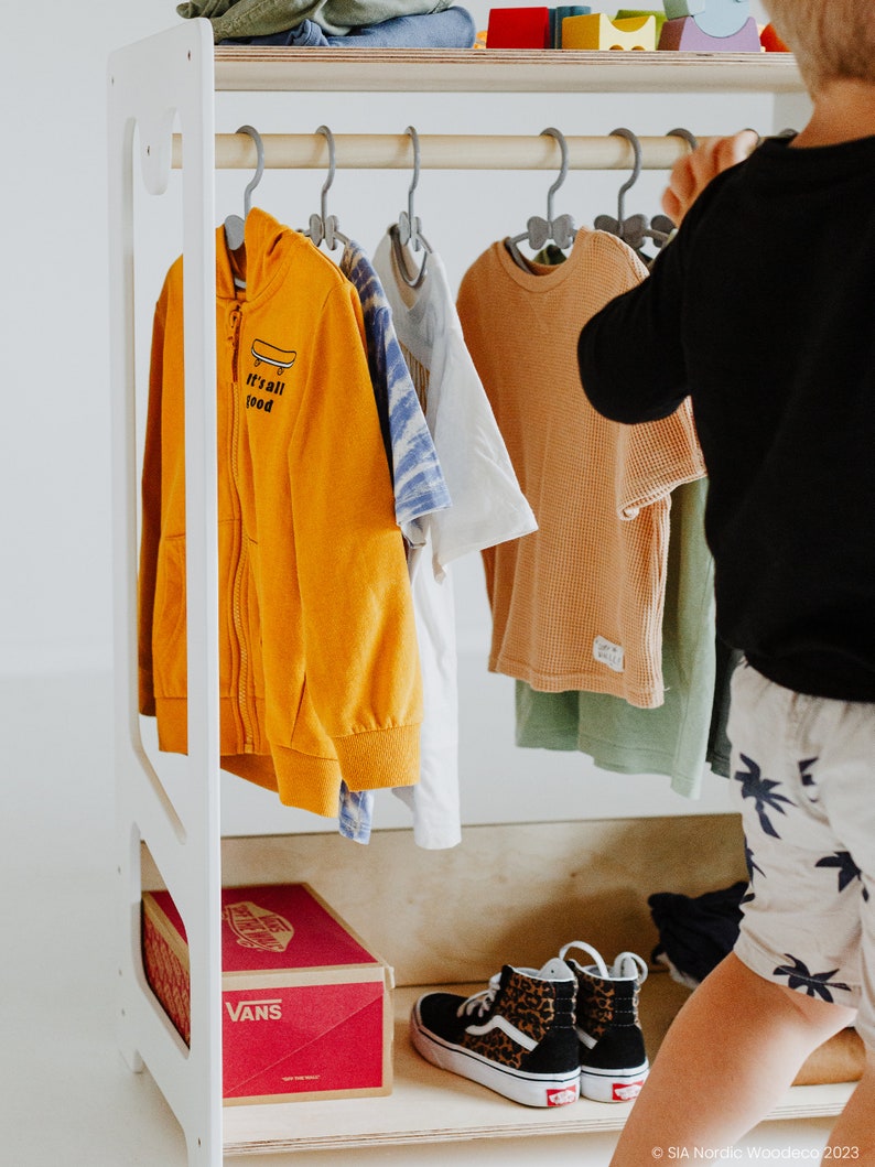 May include: A white wooden clothing rack with a shelf on top. The rack is filled with children's clothing, including a yellow hoodie with the text "it's all good", a white t-shirt, a blue and white patterned shirt, and a tan t-shirt. There is a pair of leopard print Vans shoes on the bottom shelf, along with a red box that says "Vans" on it.