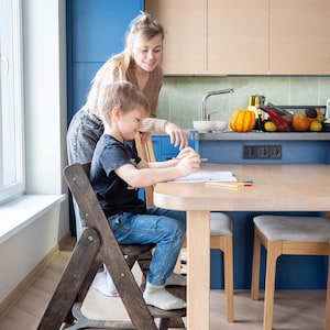 May include: A child sits at a light wood table, drawing with colored pencils, while an adult stands nearby. The child is seated on a dark wooden folding chair. The kitchen features blue cabinets and a light wood countertop.