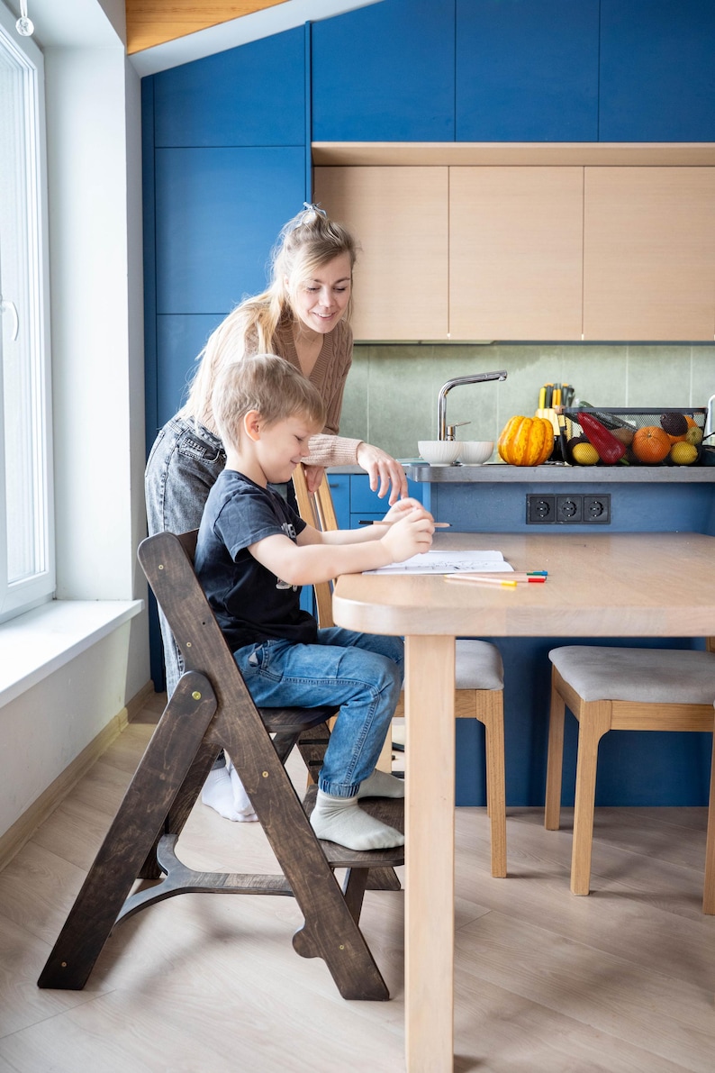 May include: A child sits at a light wood table, drawing with colored pencils, while an adult stands nearby. The child is seated on a dark wooden folding chair. The kitchen features blue cabinets and a light wood countertop.