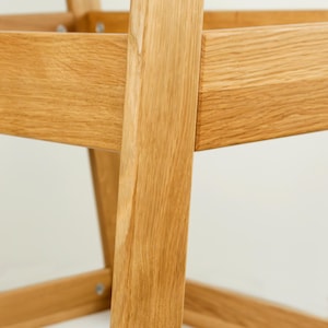 May include: Close-up of a wooden chair frame, highlighting the natural grain and texture of the light-coloured wood. The chair's construction features angled legs and a square seat frame, with visible joinery details.