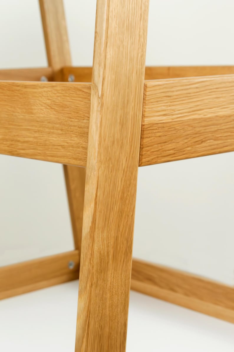 May include: Close-up of a wooden chair frame, highlighting the natural grain and texture of the light-coloured wood. The chair's construction features angled legs and a square seat frame, with visible joinery details.