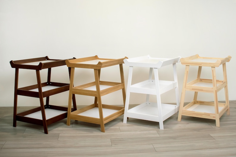 May include: Four wooden side tables in varying colors: dark brown, natural wood, white, and light wood. Each table has a tray-like top and two shelves, with a simple, modern design. The tables are suitable for various home decor styles.