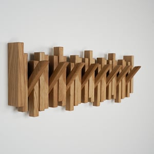 May include: Wooden wall-mounted coat rack with a modern design. The rack features multiple vertical wooden pieces of varying heights and angled hooks, all in a natural wood tone. The design is minimalist and functional.