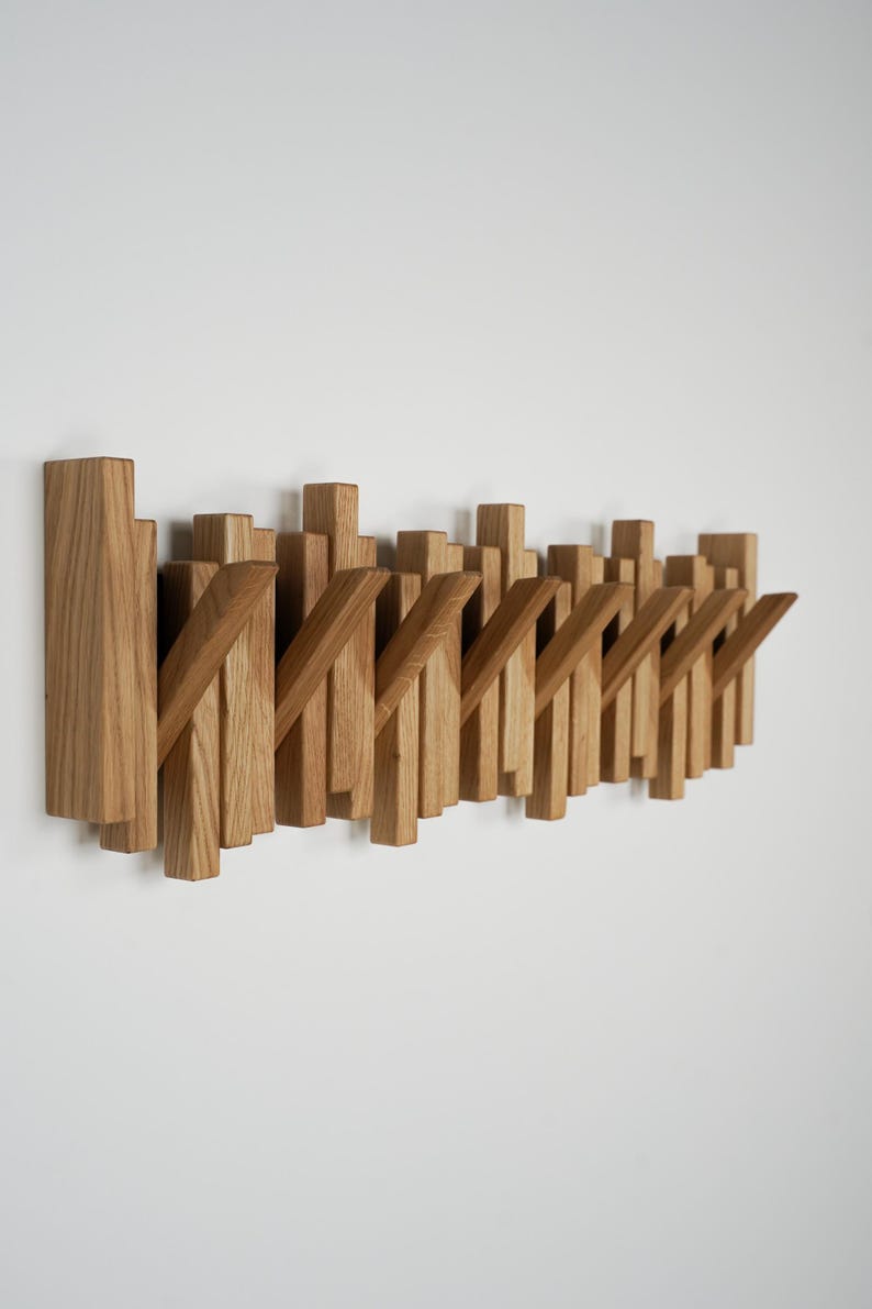 May include: Wooden wall-mounted coat rack with a modern design. The rack features multiple vertical wooden pieces of varying heights and angled hooks, all in a natural wood tone. The design is minimalist and functional.