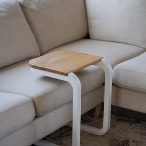 Op de afbeelding: Een moderne bijzettafel met een licht houten blad en een wit, gebogen metalen frame. De tafel is ontworpen om onder een bank te schuiven en biedt een handig oppervlak. De afmetingen zijn ongeveer 46 cm lang, 30 cm breed en 61 cm hoog.