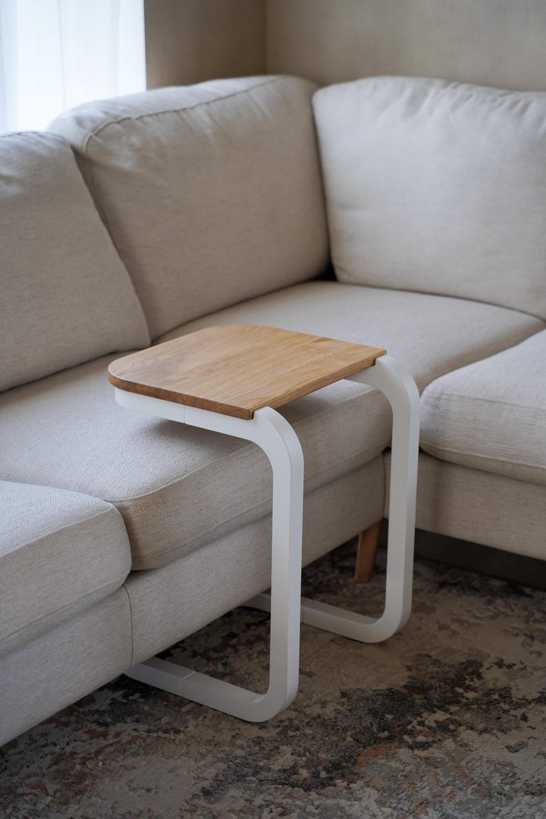Op de afbeelding: Een moderne bijzettafel met een licht houten blad en een wit, gebogen metalen frame. De tafel is ontworpen om onder een bank te schuiven en biedt een handig oppervlak. De afmetingen zijn ongeveer 46 cm lang, 30 cm breed en 61 cm hoog.