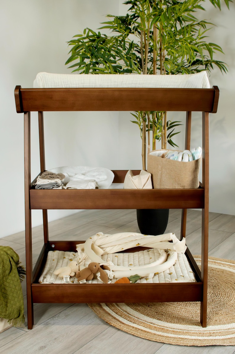 May include: A dark brown wooden changing table with three shelves. The top shelf has a white changing pad. The middle shelf holds baby supplies. The bottom shelf has a quilted pad and toys. A jute rug is on the floor.