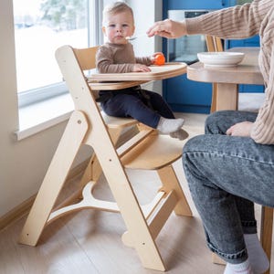 May include: A light-colored wooden high chair with a tray, designed for a baby. The chair has a safety harness and is positioned near a window. A person is feeding the baby with a spoon.