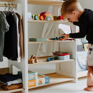 May include: White wooden children's clothing rack and bookshelf with a rainbow wooden toy, books, and toys on the shelves. A young child is standing on the bottom shelf, reaching for a toy.