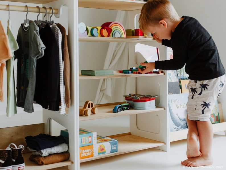 May include: White wooden children's clothing rack and bookshelf with a rainbow wooden toy, books, and toys on the shelves. A young child is standing on the bottom shelf, reaching for a toy.