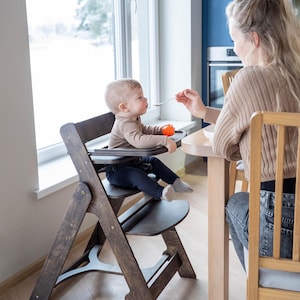 May include: A wooden high chair with a dark brown finish. The chair has a tray and footrest. A baby is seated in the chair, being fed with a spoon. The background includes a window and a person.