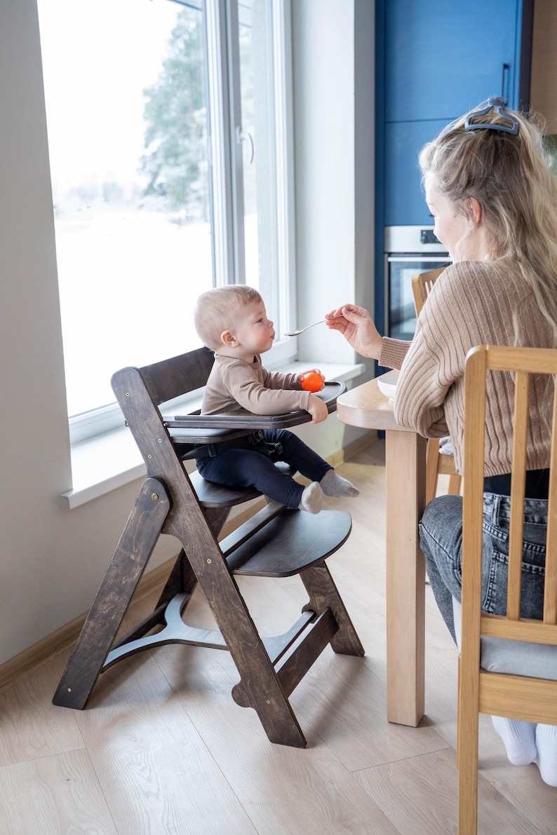 May include: A wooden high chair with a dark brown finish. The chair has a tray and footrest. A baby is seated in the chair, being fed with a spoon. The background includes a window and a person.