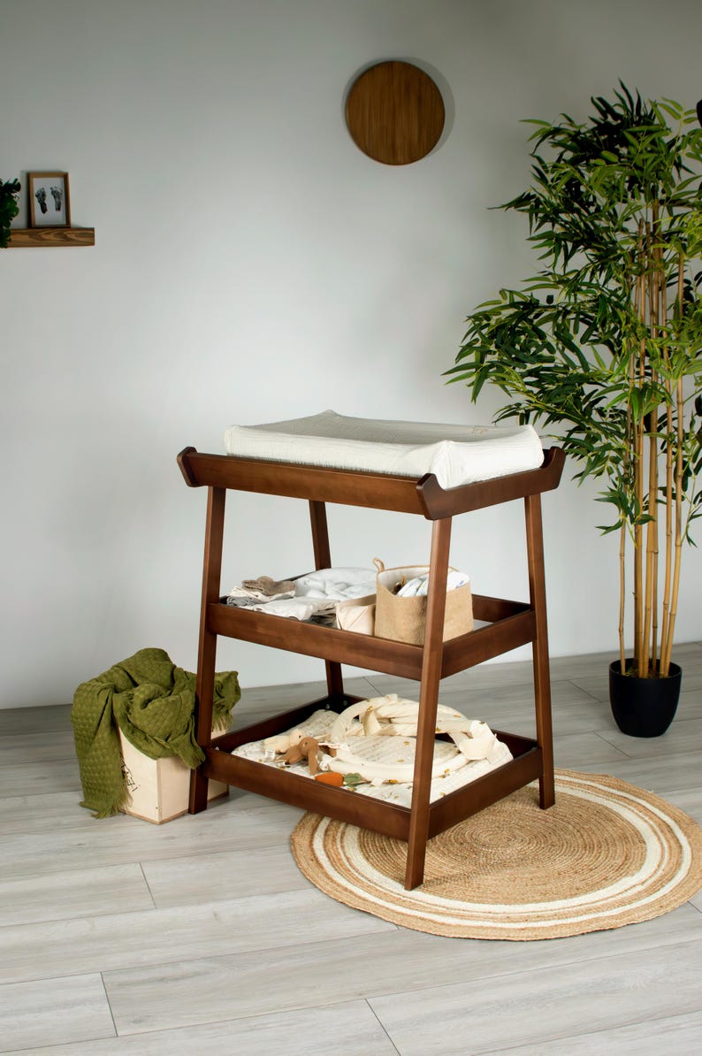May include: A wooden changing table with three shelves. The top shelf has a white changing pad. The lower shelves hold baby supplies. A green blanket and a wooden box are on the floor. A round rug is beneath the table.