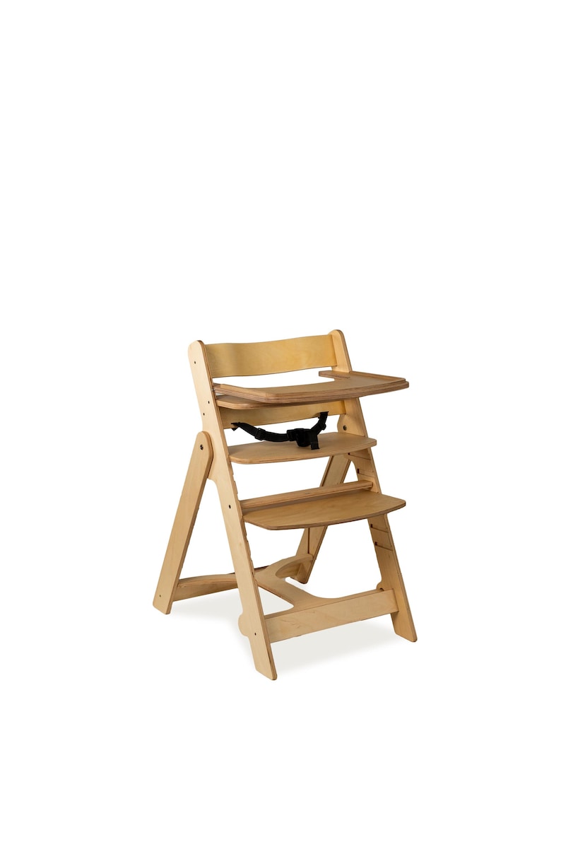 May include: A wooden high chair with a tray and footrest. The chair is made of light-colored wood and has a black safety strap. The design is simple and functional, suitable for a baby or toddler.