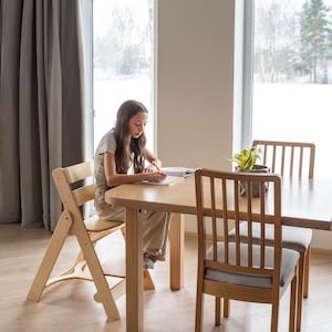 May include: A light wood dining table and chairs set. The table has a rounded edge and is surrounded by two chairs with gray cushions and a folding chair. A person is seated at the table, writing.