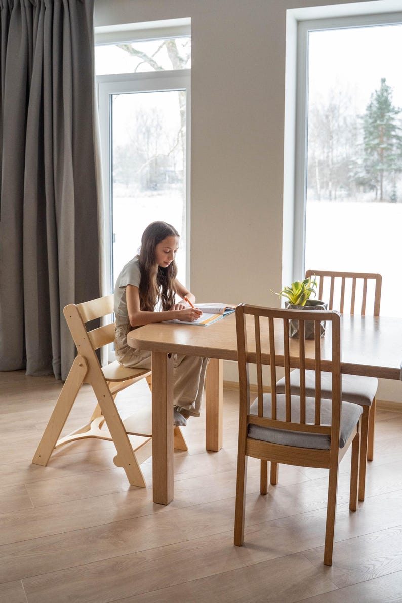 May include: A light wood dining table and chairs set. The table has a rounded edge and is surrounded by two chairs with gray cushions and a folding chair. A person is seated at the table, writing.