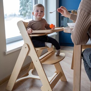 May include: A light-colored wooden high chair with a tray, holding a baby wearing a brown shirt and black pants. The baby is holding an orange ball. A person is feeding the baby with a spoon.