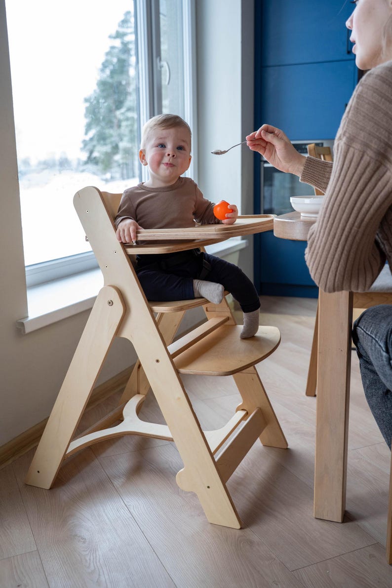 May include: A light-colored wooden high chair with a tray, holding a baby wearing a brown shirt and black pants. The baby is holding an orange ball. A person is feeding the baby with a spoon.