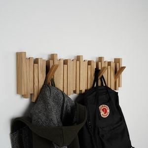 May include: A wooden wall-mounted coat rack with a unique design. The rack features multiple vertical wooden pieces of varying heights. A black backpack and a dark green coat with a hood are hanging from the rack.