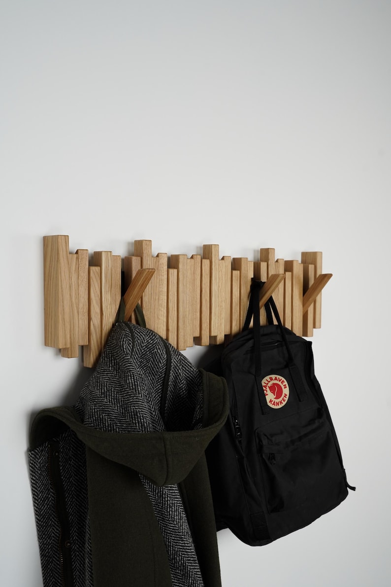 May include: A wooden wall-mounted coat rack with a unique design. The rack features multiple vertical wooden pieces of varying heights. A black backpack and a dark green coat with a hood are hanging from the rack.